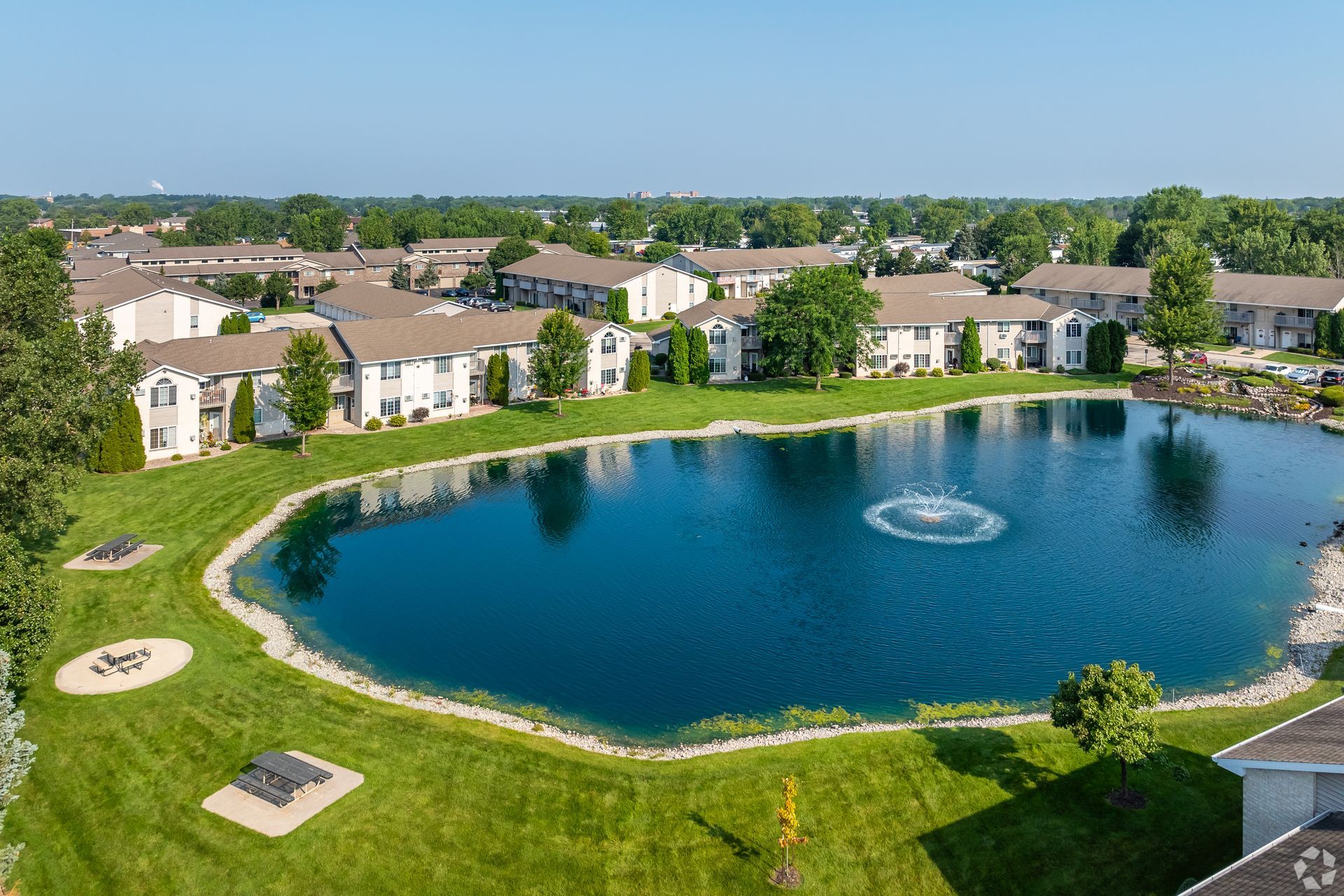 Aerial view of apartment homes around a landscaped pond with a fountain and green lawns.