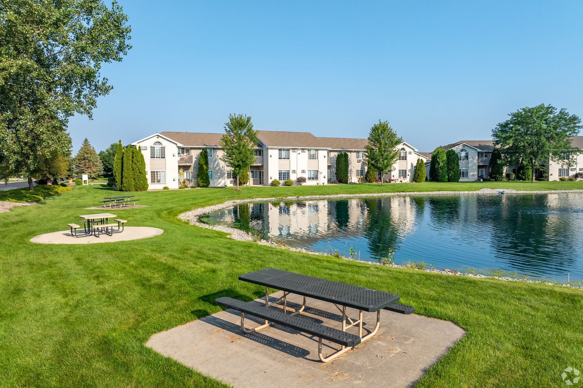 Apartment buildings by a pond with picnic tables and green lawns under a clear blue sky