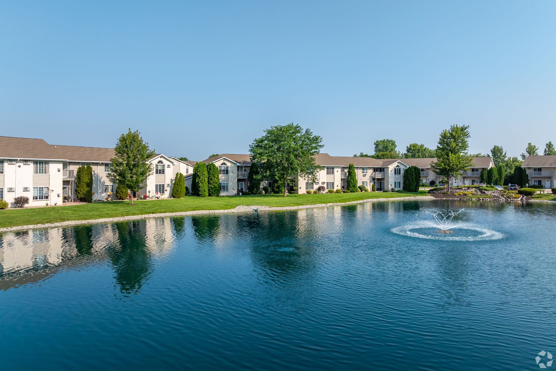 Apartment buildings reflected in a calm pond with a fountain under a clear blue sky