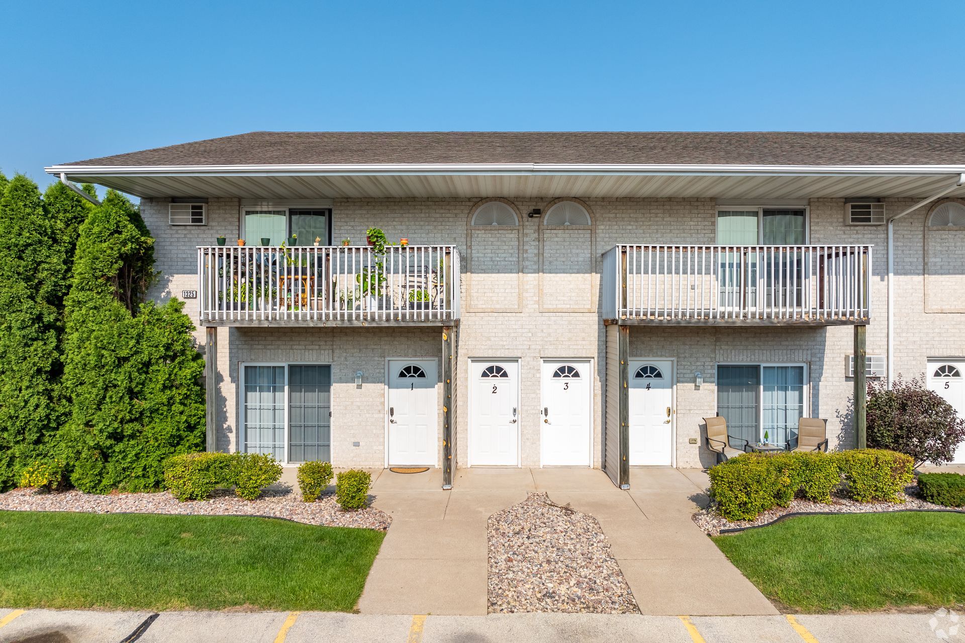 Two-story beige apartment building with balconies, white doors, and a central walkway surrounded by landscaping