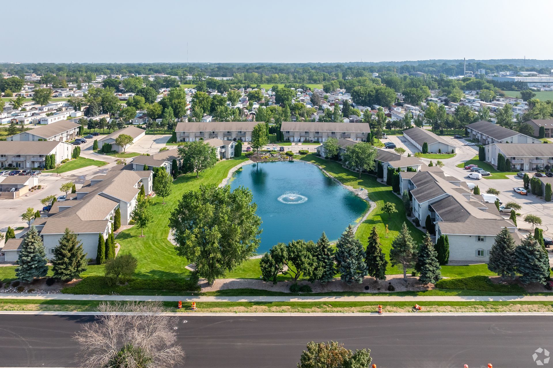 Aerial view of an apartment complex with a central pond and fountain, surrounded by trees and roads