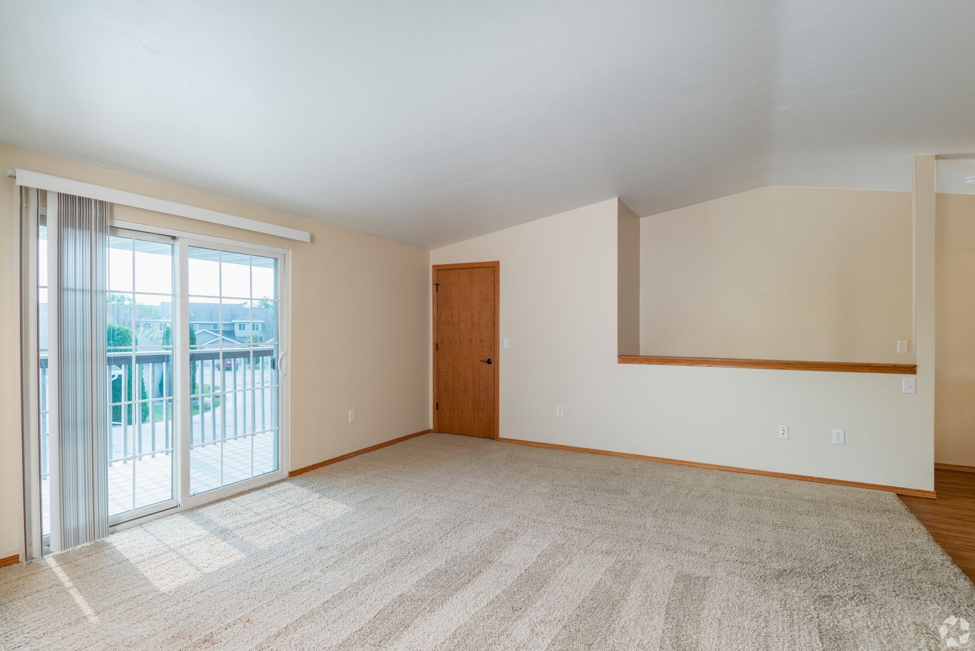 Empty beige living room with carpet, sliding glass door, and wooden door near a half-wall opening