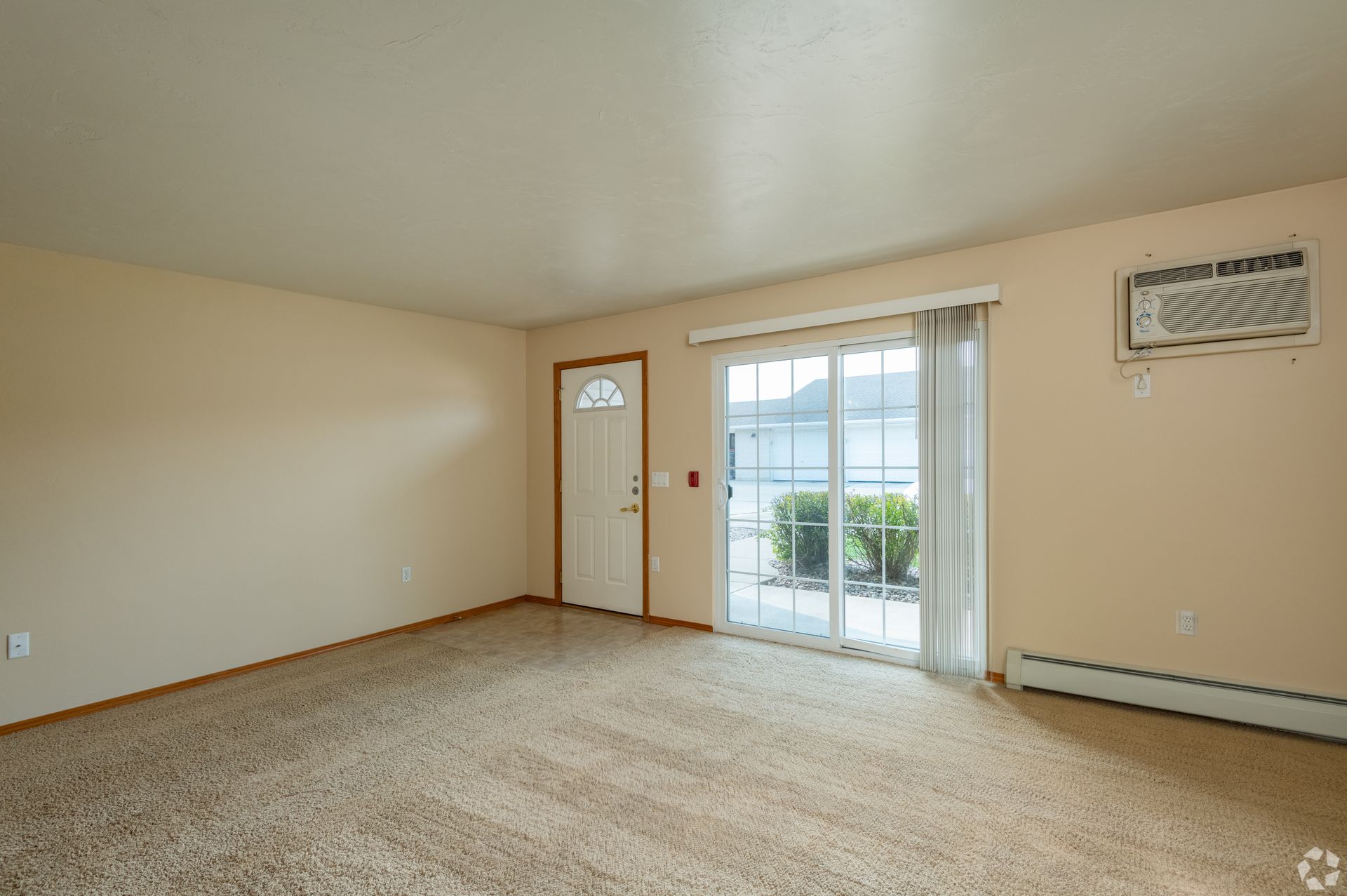 Empty beige living room with carpet, door, sliding glass doors, and wall air conditioner