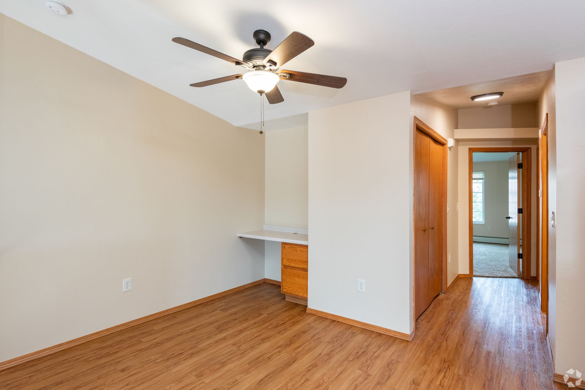 Empty apartment room with wood floors, ceiling fan, and hallway leading to kitchen area.