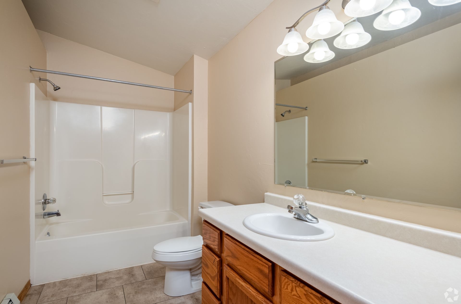 Bathroom with white tub-shower, toilet, and wooden vanity with large mirror and lights.