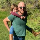 Man with sunglasses giving a piggyback ride to a child outdoors; both smiling.