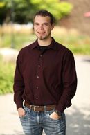 Man smiles, hands in pockets, wearing a burgundy shirt, jeans, and brown belt, outdoors.