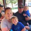 Family smiling together on a train; parents hold two young children. Outdoors setting, sunny.