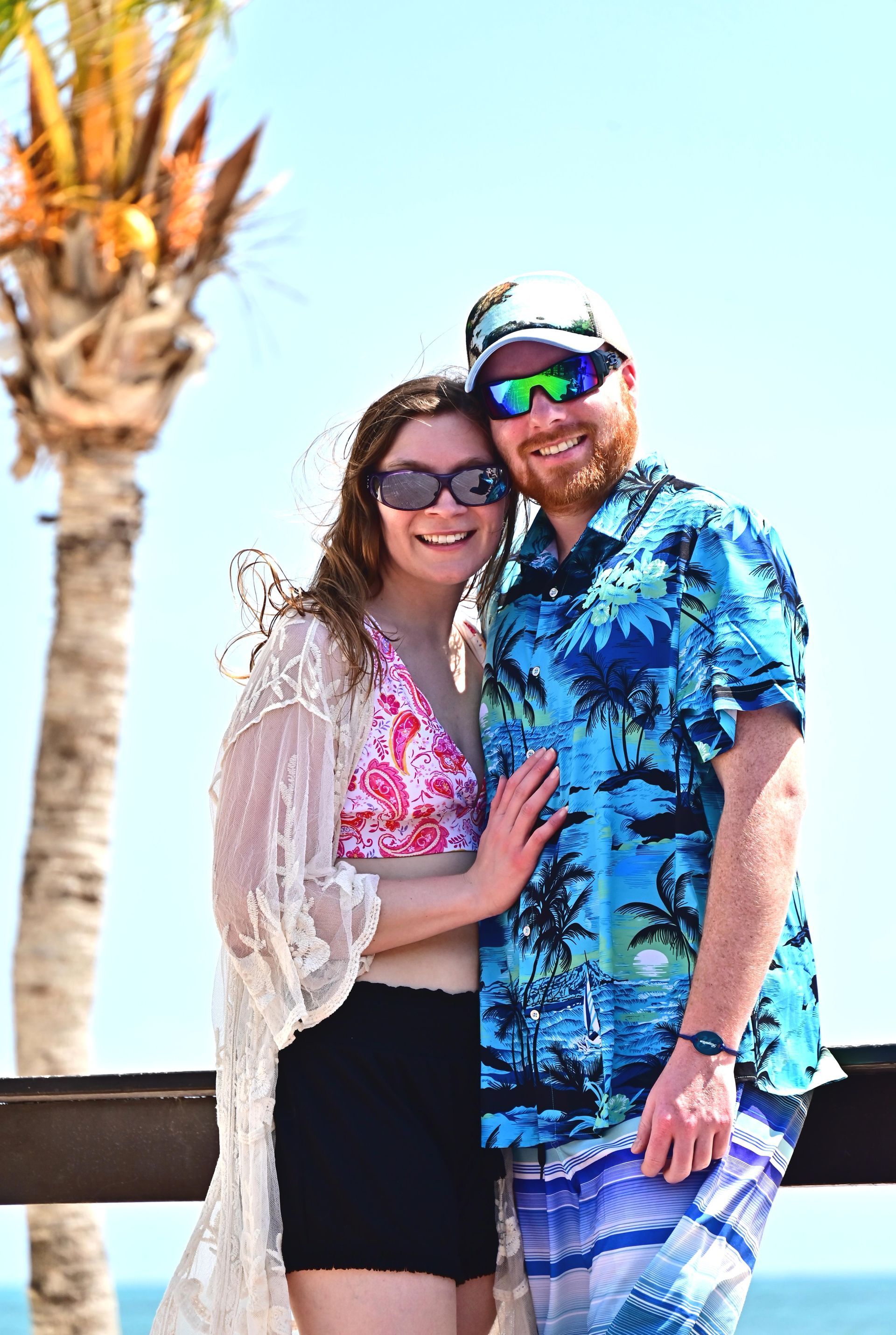 Couple smiling, embracing outdoors near beach. Woman in bikini and lace cover-up. Man in Hawaiian shirt and sunglasses.