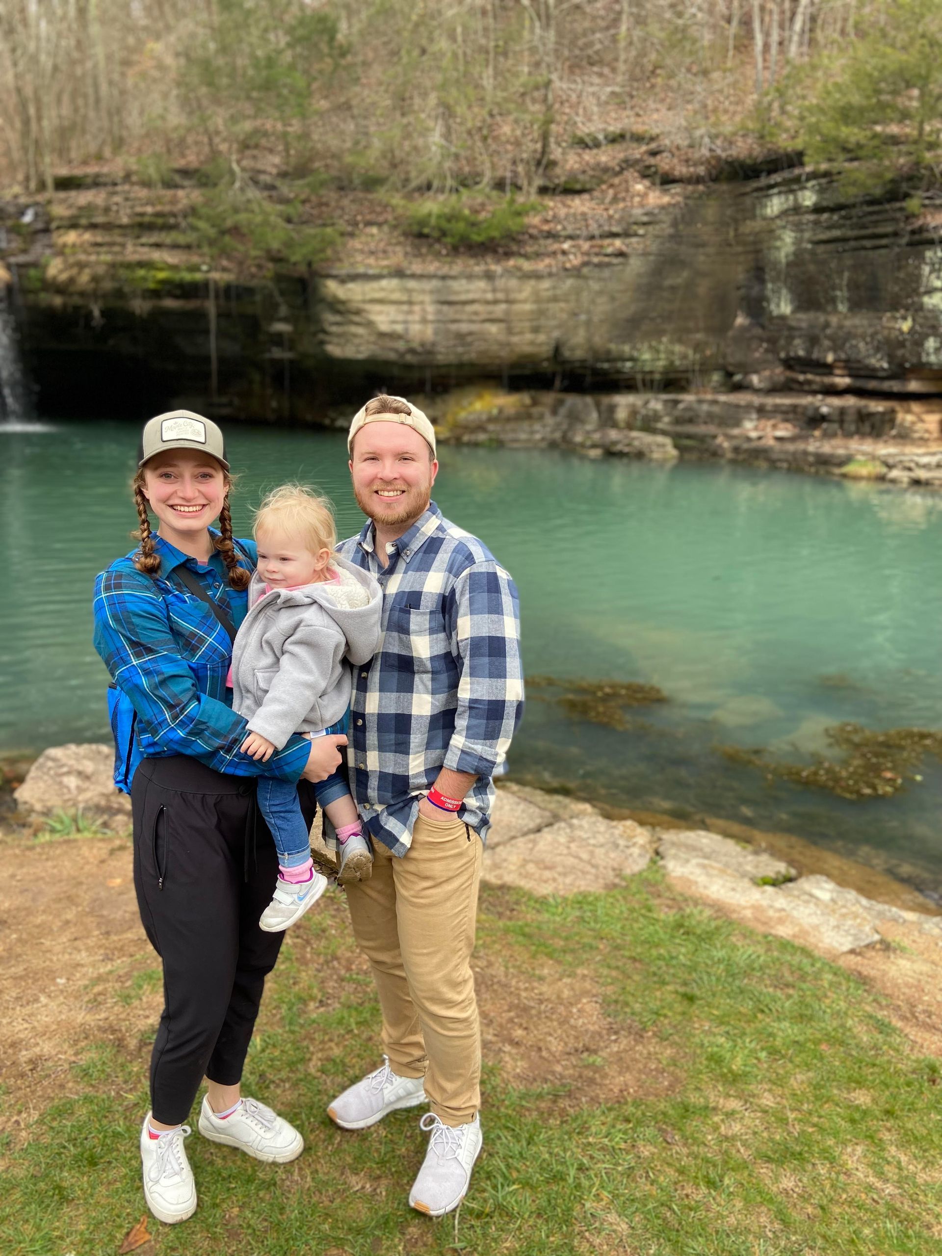 Family of three at a turquoise-watered waterfall. Woman holds child, man stands next to them. Outdoors.