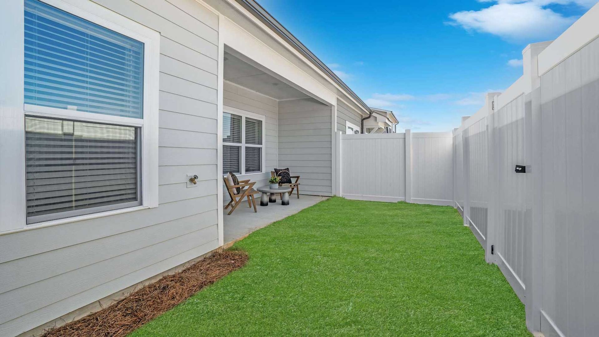 Narrow backyard with green lawn, white fence, and patio beside a light-gray house under a blue sky