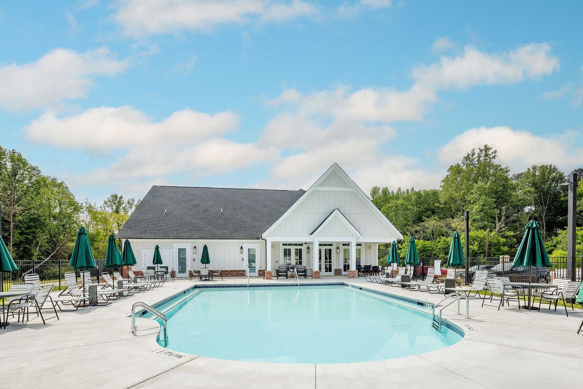 Outdoor resort pool with lounge chairs, closed umbrellas, and a white clubhouse under a blue sky