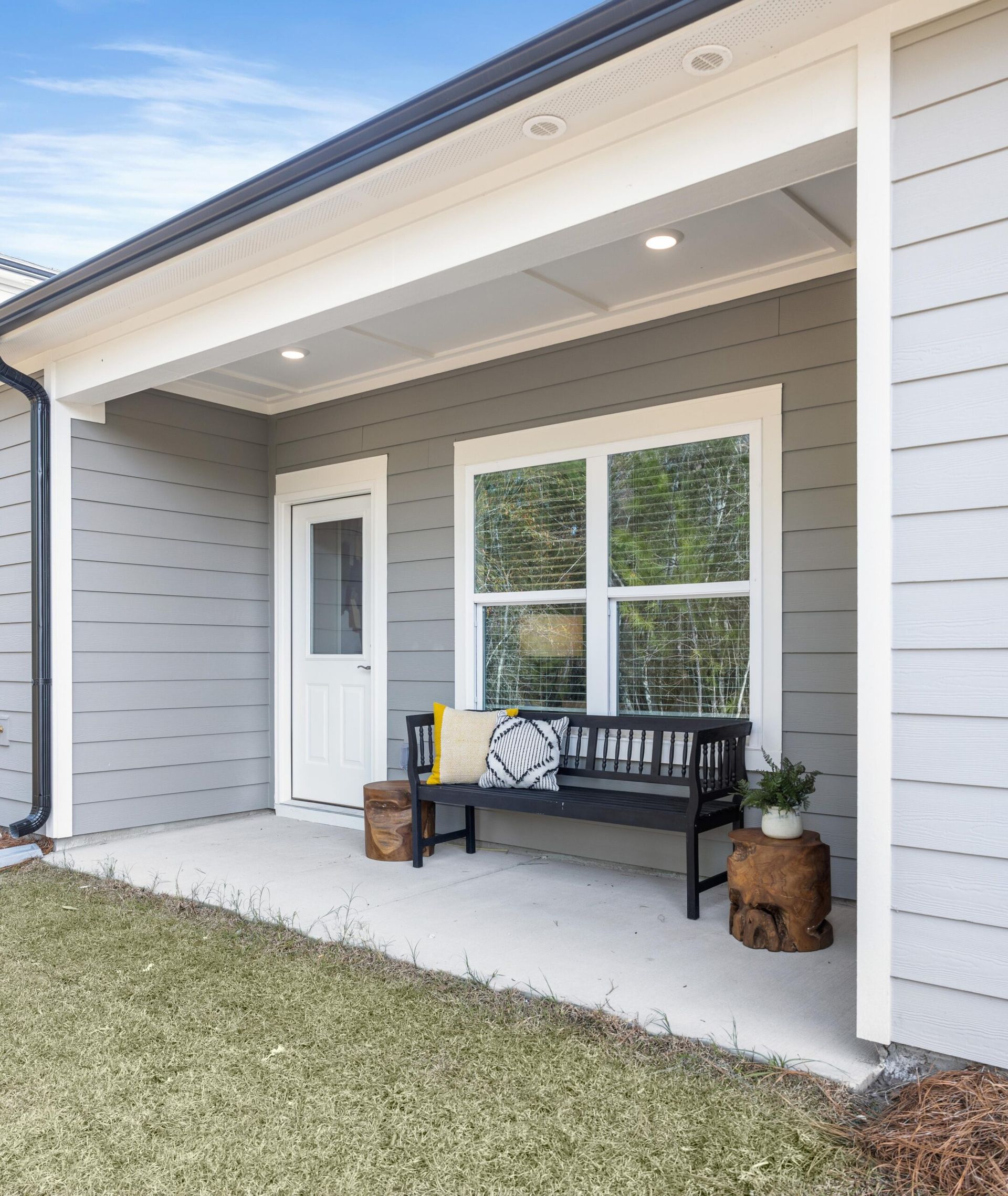 Covered front porch with gray siding, white door, bench, and potted plants in a modern home.