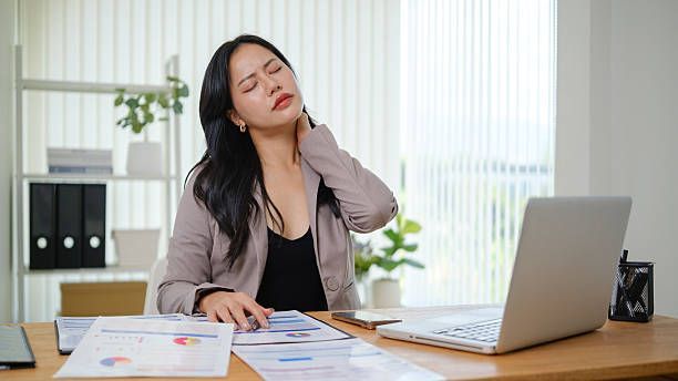Office worker rubbing neck beside laptop at desk, looking uncomfortable with papers and charts nearby