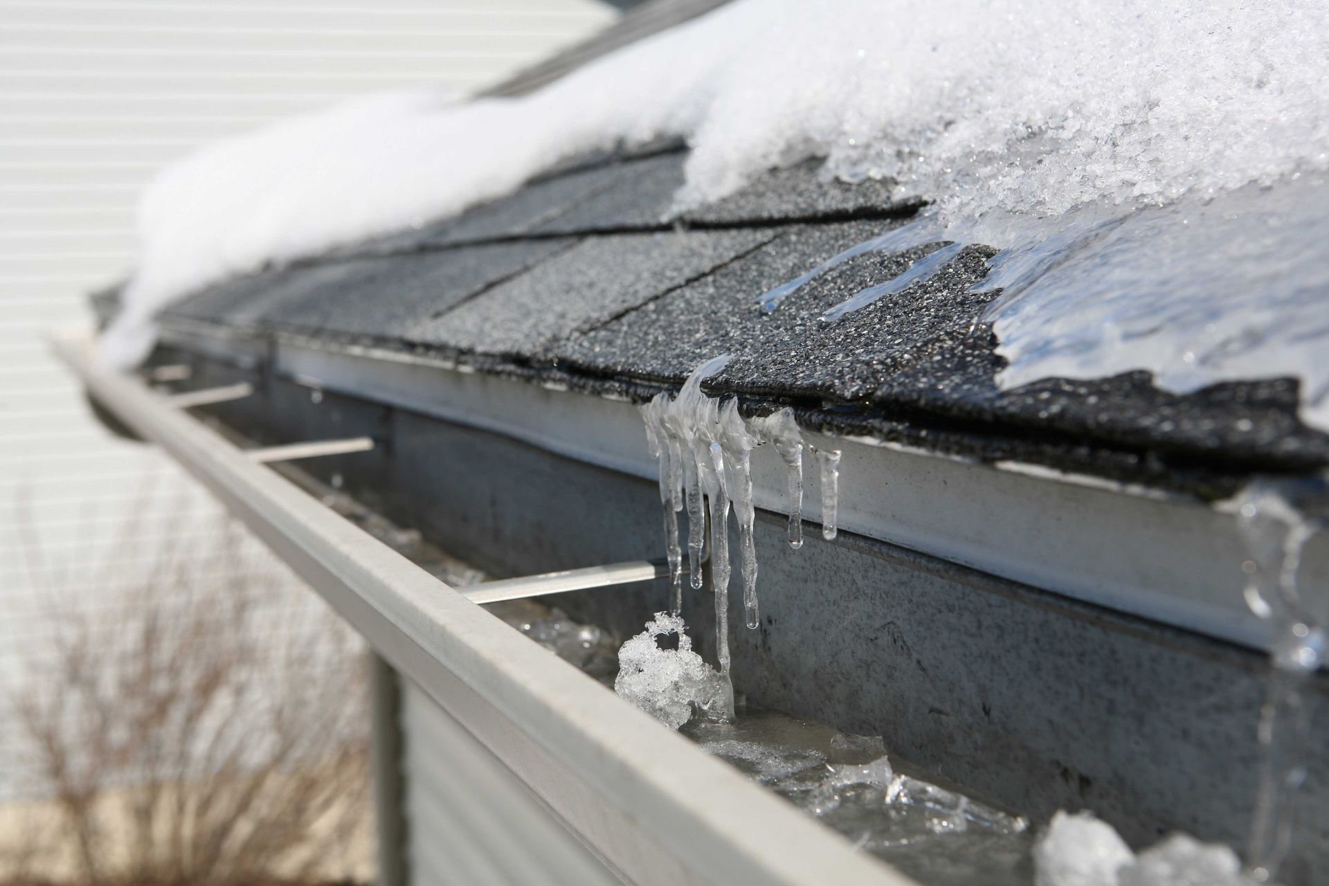 Gros plan sur une gouttière de toit remplie de neige et de glace fondantes, avec des stalactites de glace suspendues aux bardeaux au-dessus.