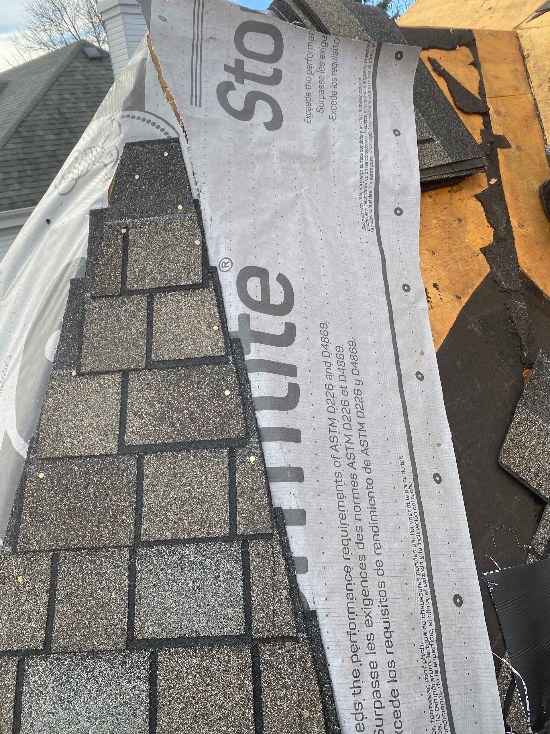Un toit en construction montre des bardeaux d'asphalte recouvrant une sous-couche synthétique grise sur un revêtement en bois apparent.
