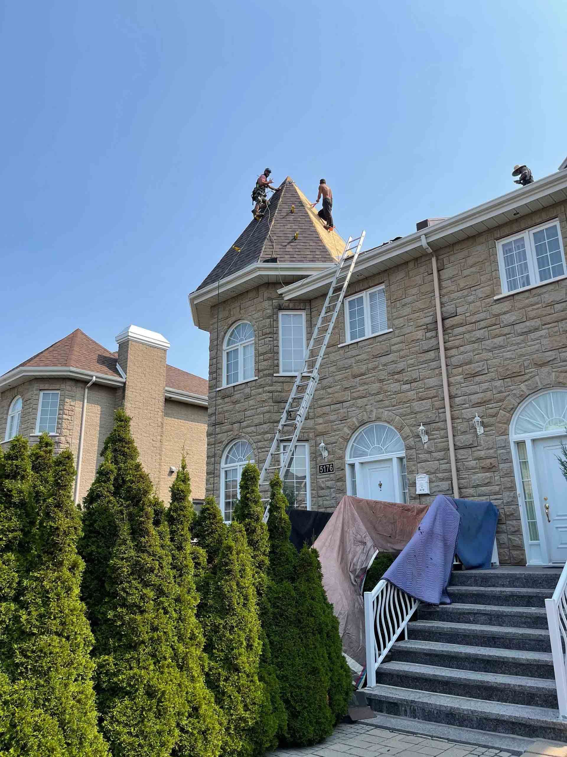Stone house with roof repair crew on scaffolding beside outdoor stairs and trimmed hedges under a blue sky