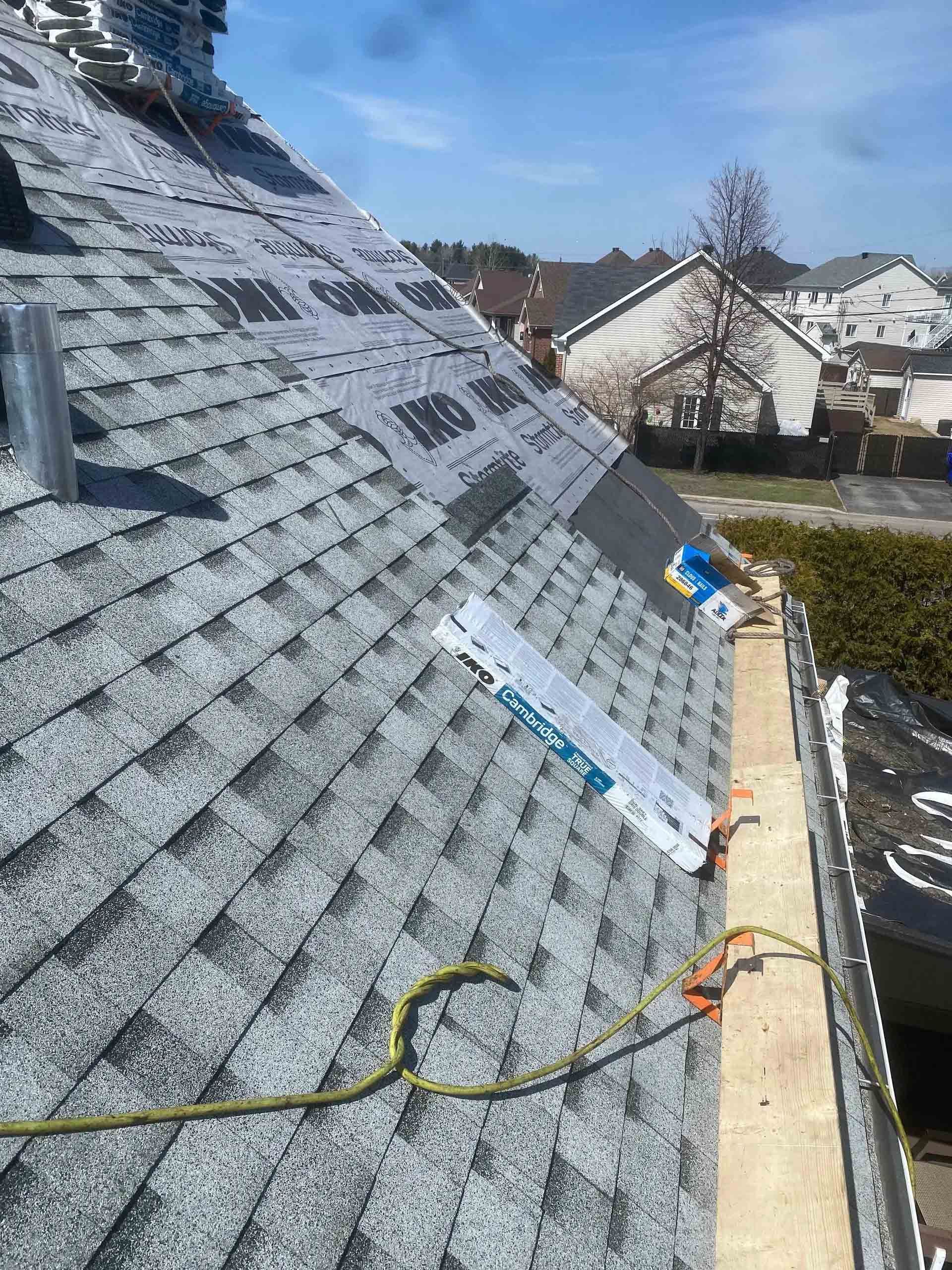 Roof under repair with gray shingles, safety harness lines, and neighboring houses in the background