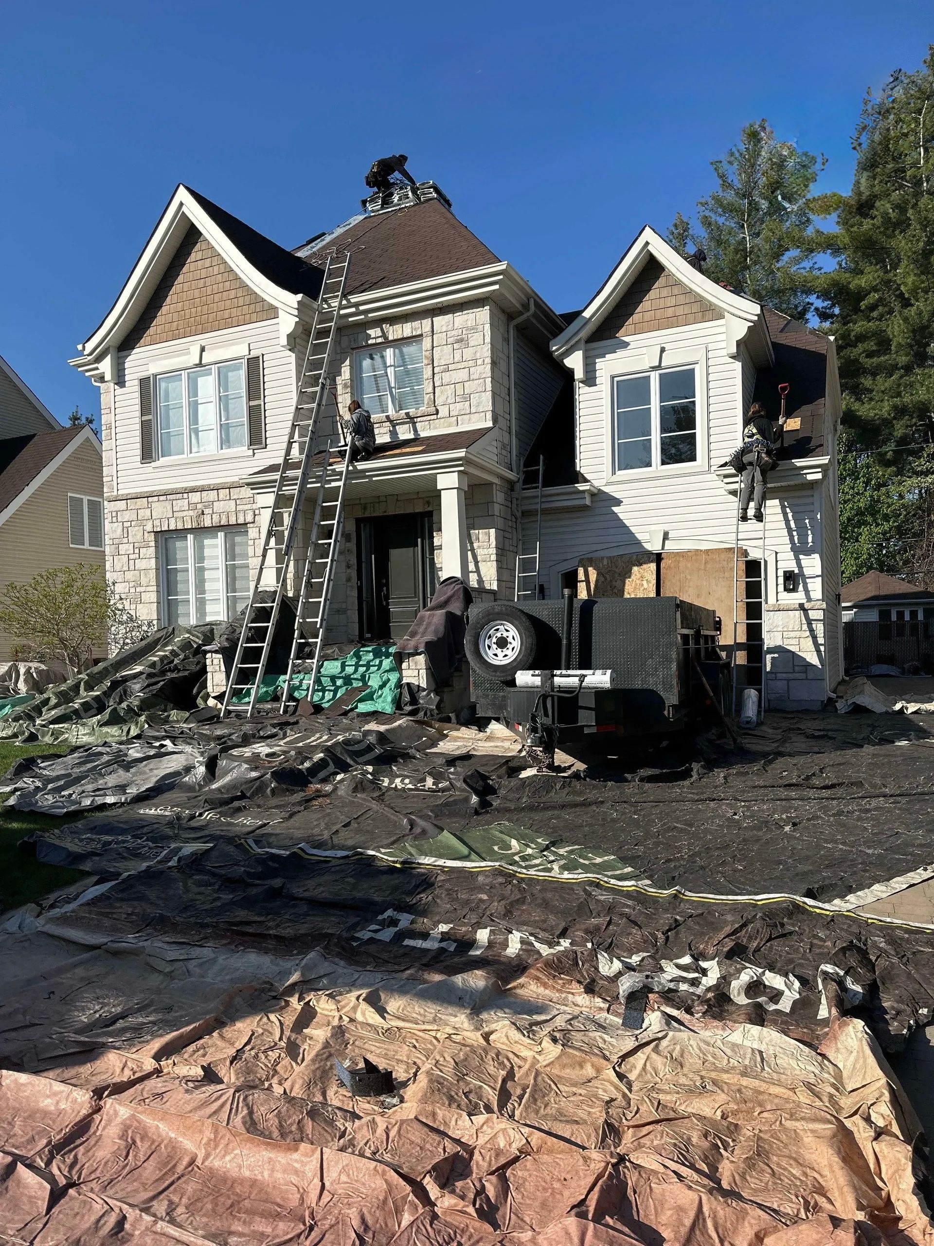 Une grande maison à deux étages en rénovation, avec des échafaudages, des débris dans la cour et une remorque utilitaire sombre garée devant.
