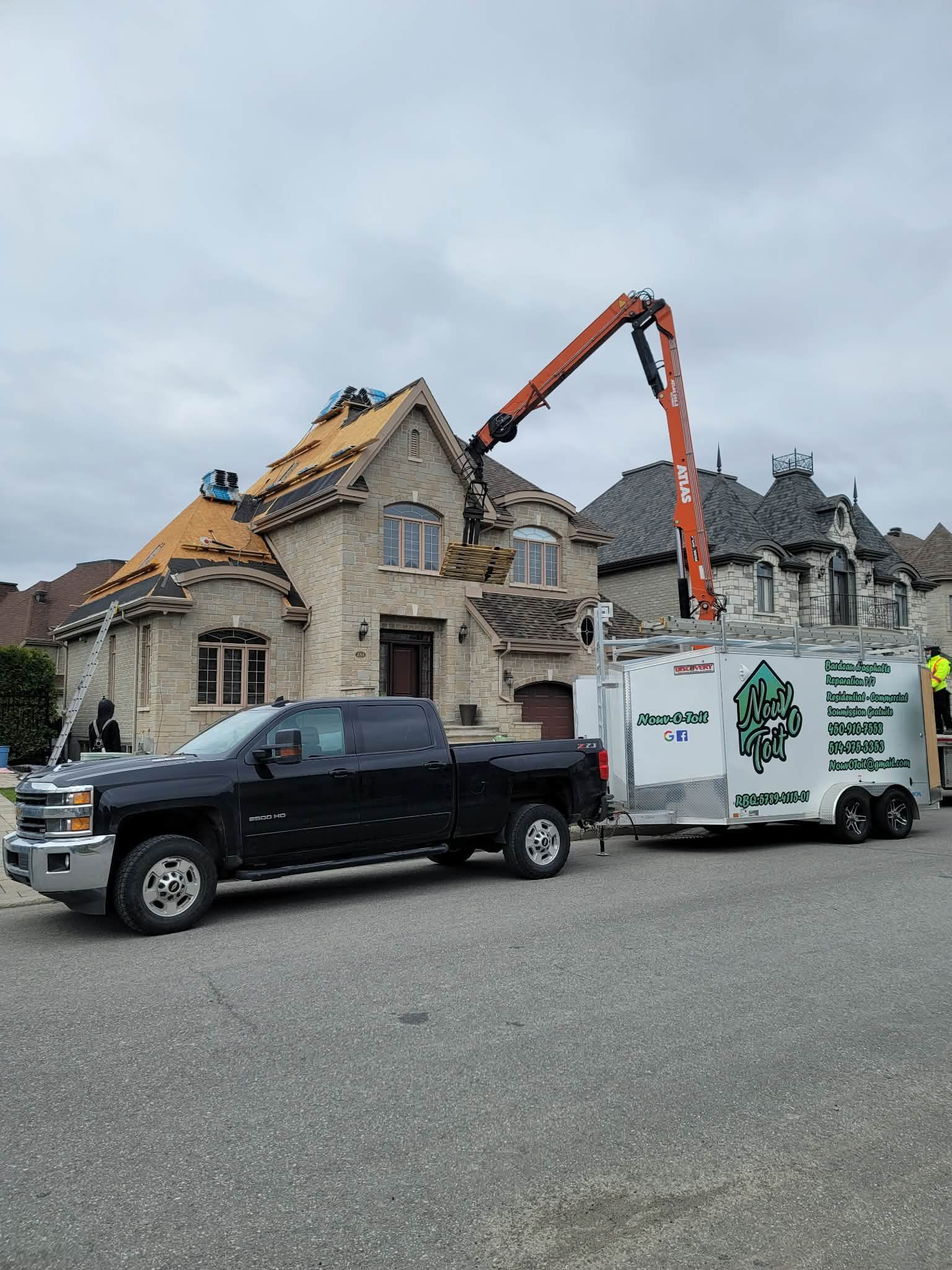 Une camionnette noire remorque une remorque de chantier dont la longue flèche de grue orange s'étend vers le toit d'une maison en pierre.