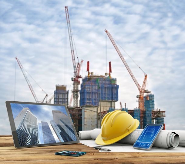 Cantiere edile con gru, computer portatile, elmetto, smartphone e progetti arrotolati su una superficie di legno.