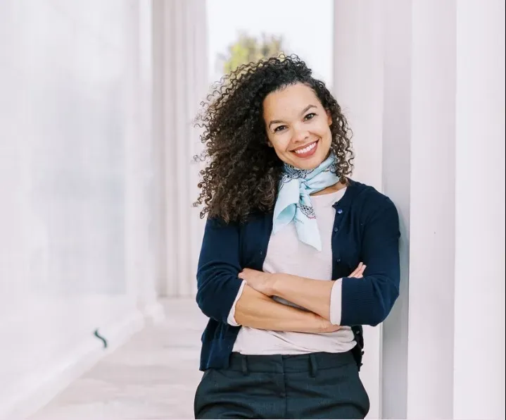 Smiling person leaning against white columns, wearing a navy blazer and scarf, with arms crossed.