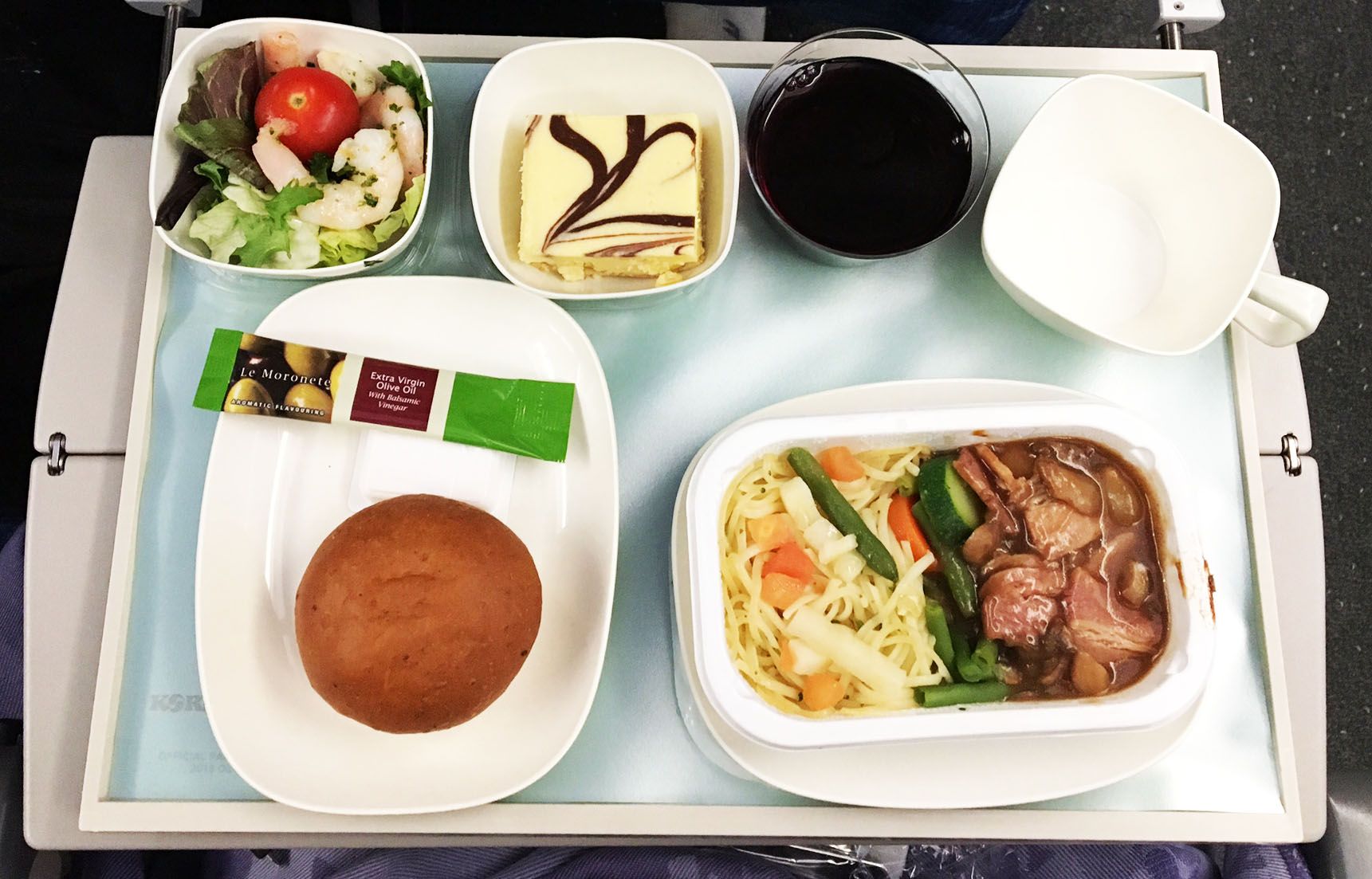 Airline meal tray with salad, dessert, bread roll, coffee, water, and a plated main dish
