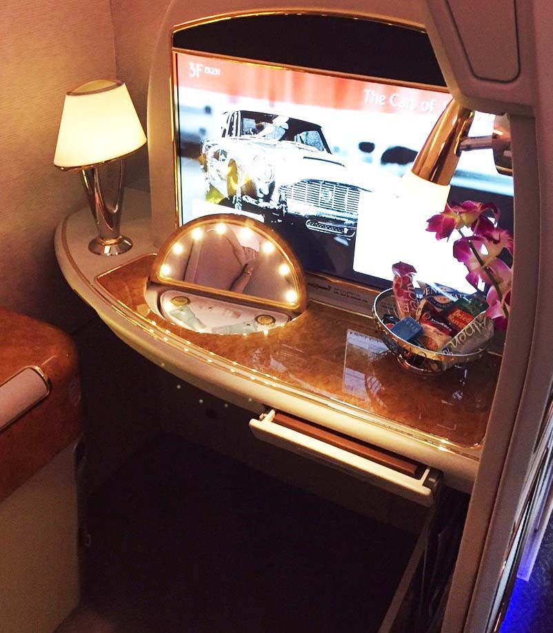 Airplane first-class seat with a TV, lamp, flower vase, and lit vanity table on a wooden side console