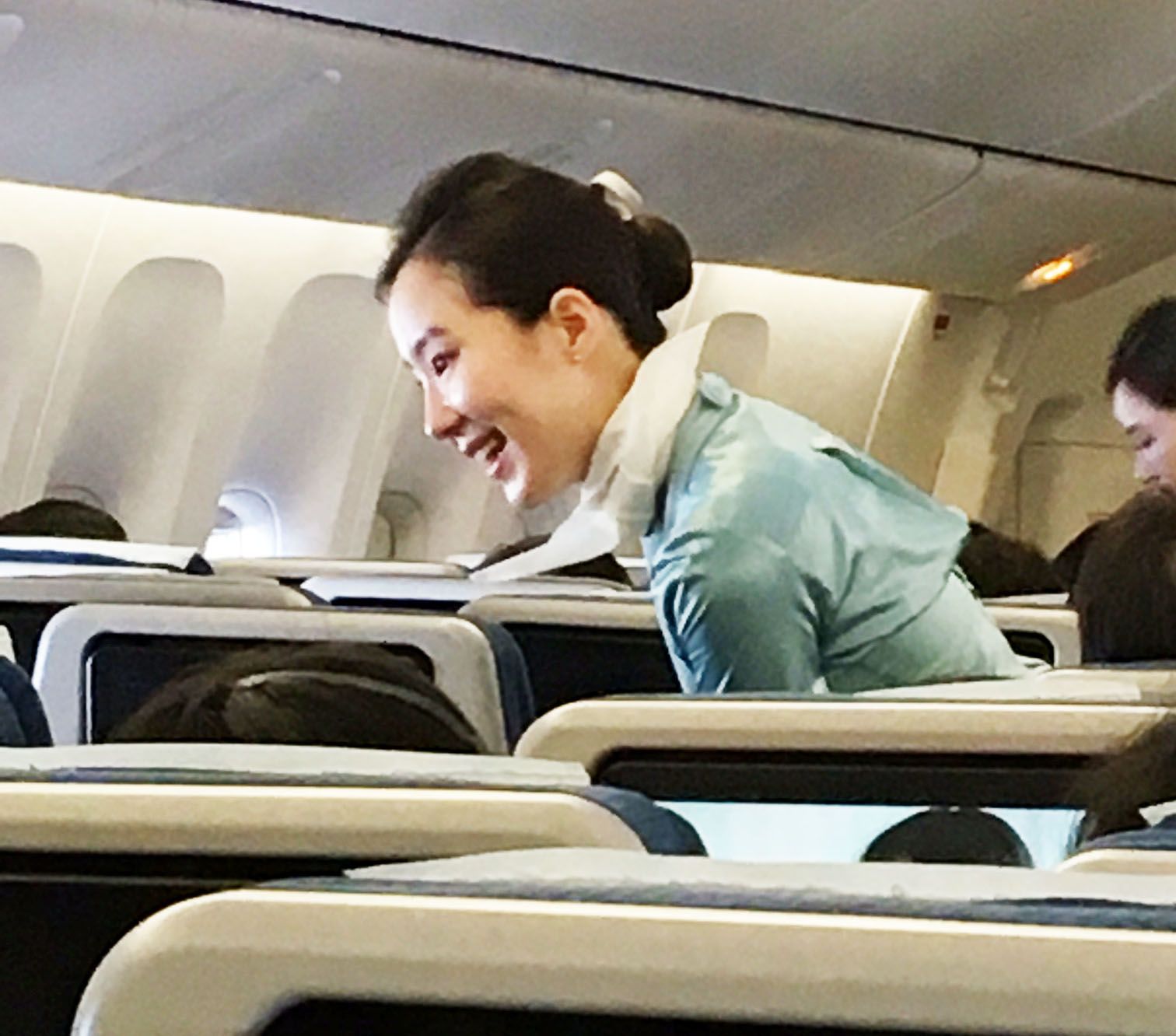 Smiling woman in a teal jacket leaning over an airplane seat on a flight