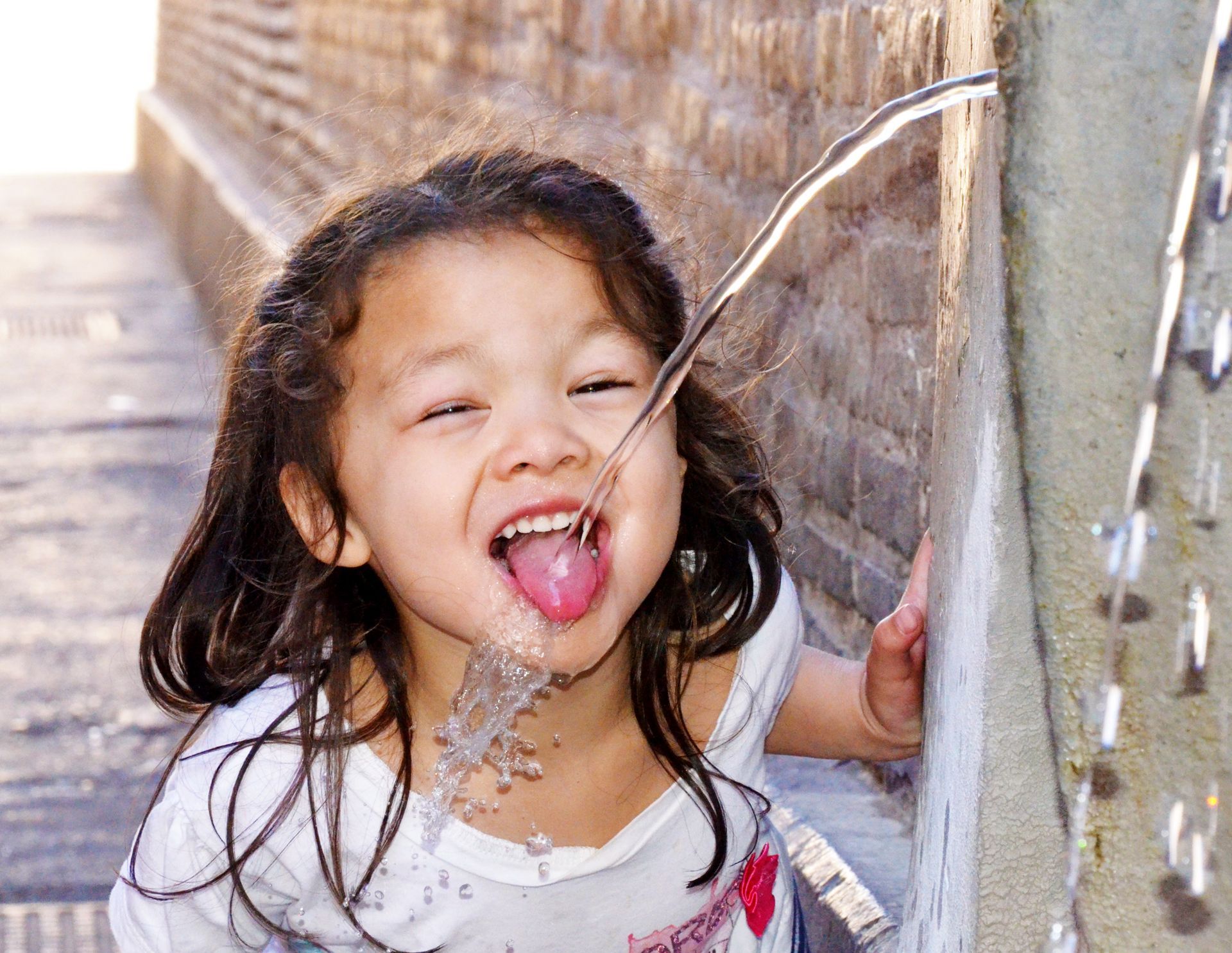 Child laughing in a water spray beside a wooden fence, wet hair and tongue out.