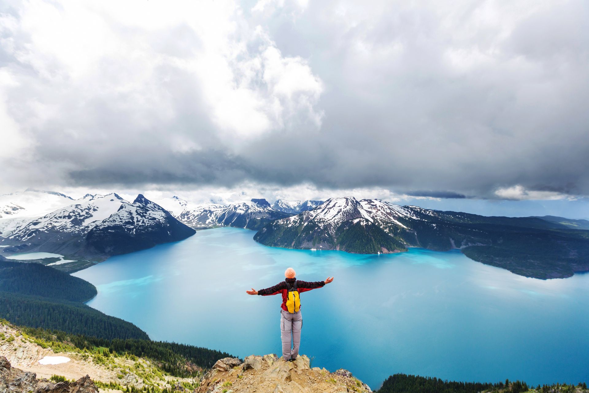 Person standing on a cliff overlooking a turquoise mountain lake and snow-covered peaks under cloudy skies