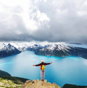 Person with arms outstretched overlooking a turquoise mountain lake under cloudy skies