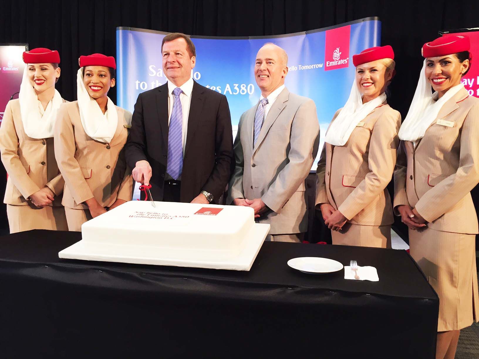 Group posing at a cake-cutting event with a large cake, table, and blue banner backdrop