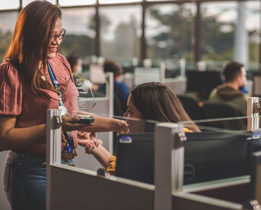 Coworkers talking across office cubicles, one handing over a small item in a bright workspace