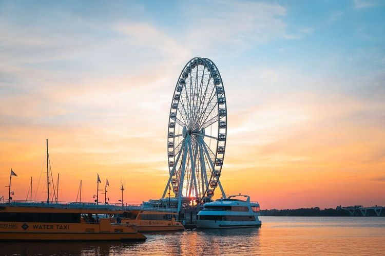 Ferris wheel at sunset over calm water with boats and a colorful sky