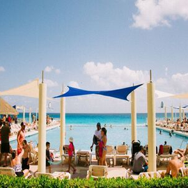 Beachside resort pool with lounge chairs, blue shade sails, and people relaxing under a bright sky