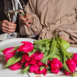 Two people clink champagne glasses beside a bouquet of pink tulips on a dining table.