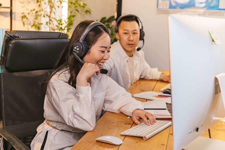 Two office workers wearing headsets at a desk, using a computer in a bright office