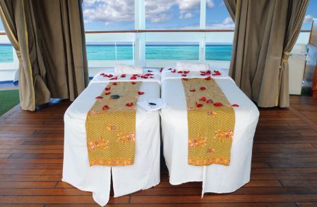 Two spa massage tables by a window overlooking the ocean, with white linens and gold runners.