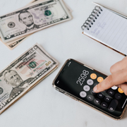 Calculator and dollar bills on a white desk, with a hand entering a number on a smartphone calculator.