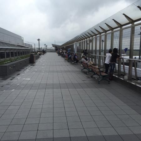 Long tiled walkway with benches and people beside a fenced waterfront under a cloudy sky.