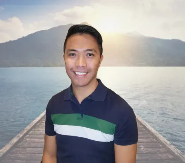 Smiling person in navy polo standing on a dock with calm water and mountains at sunset
