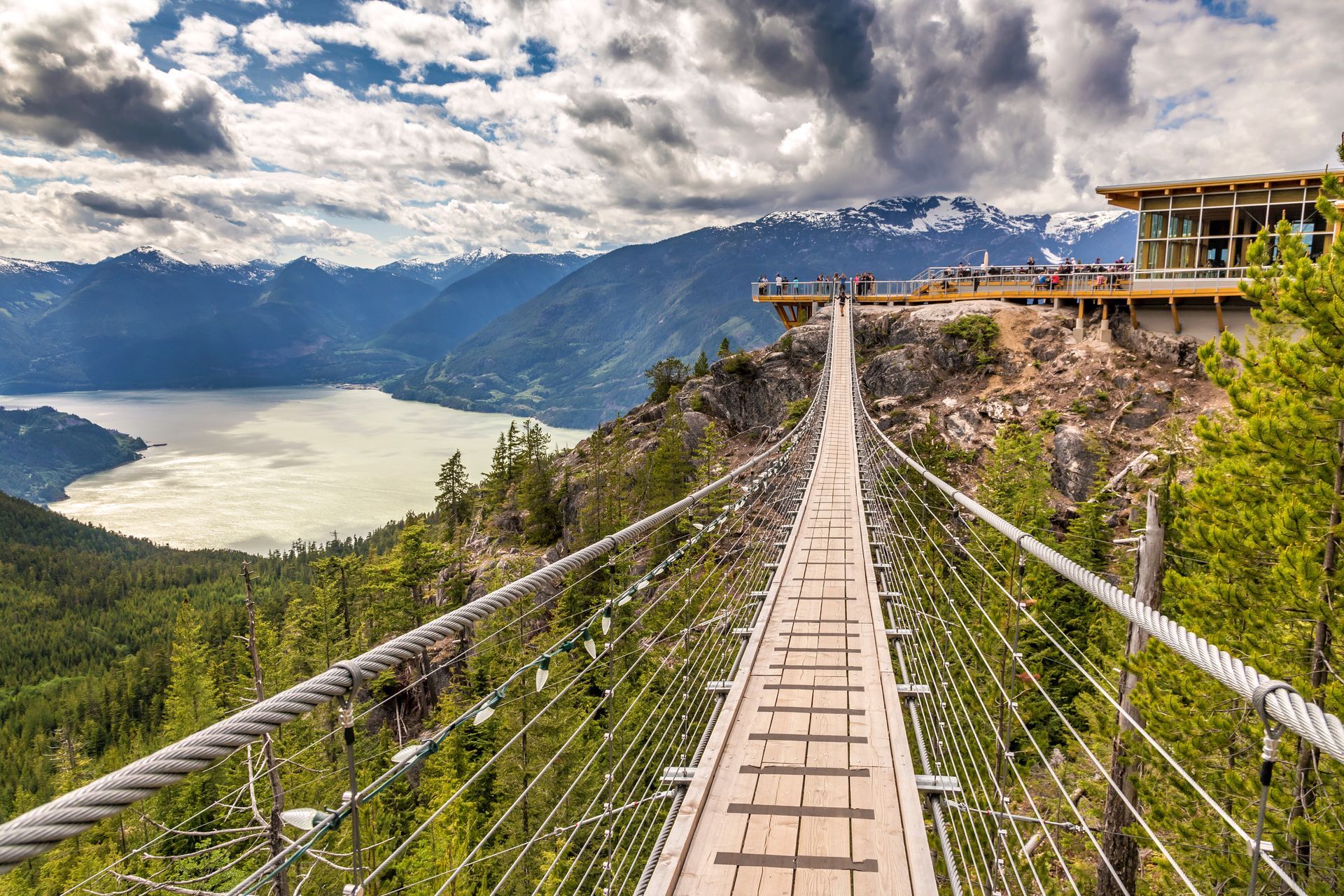 Suspension bridge over a forested valley and lake, with mountains and a cloudy sky in the background
