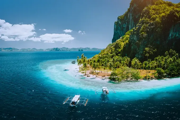 Tropical island with turquoise water, white sandy beach, steep green cliffs, and small boats offshore