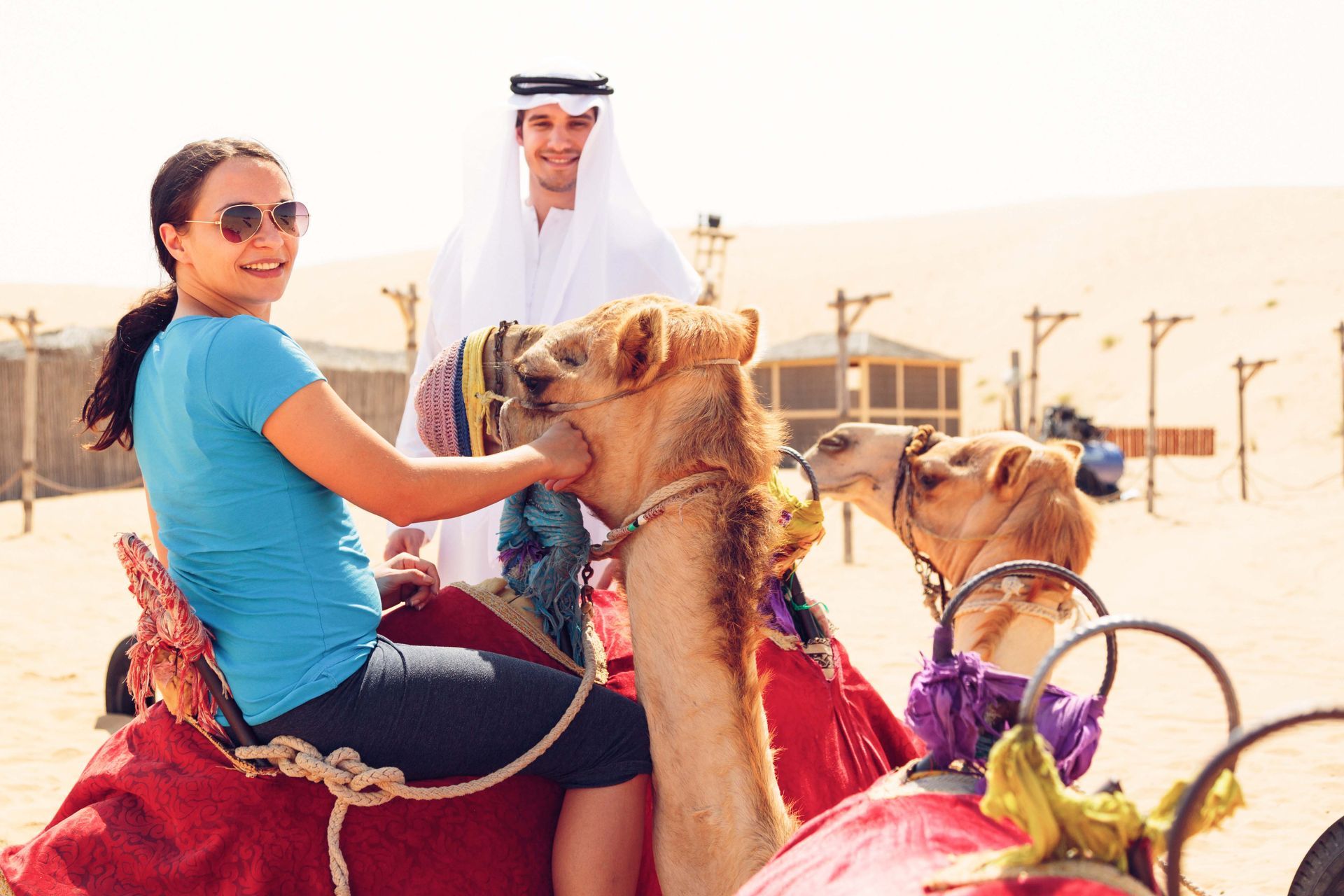 Woman riding a camel in a desert tour, with a man in traditional dress standing nearby