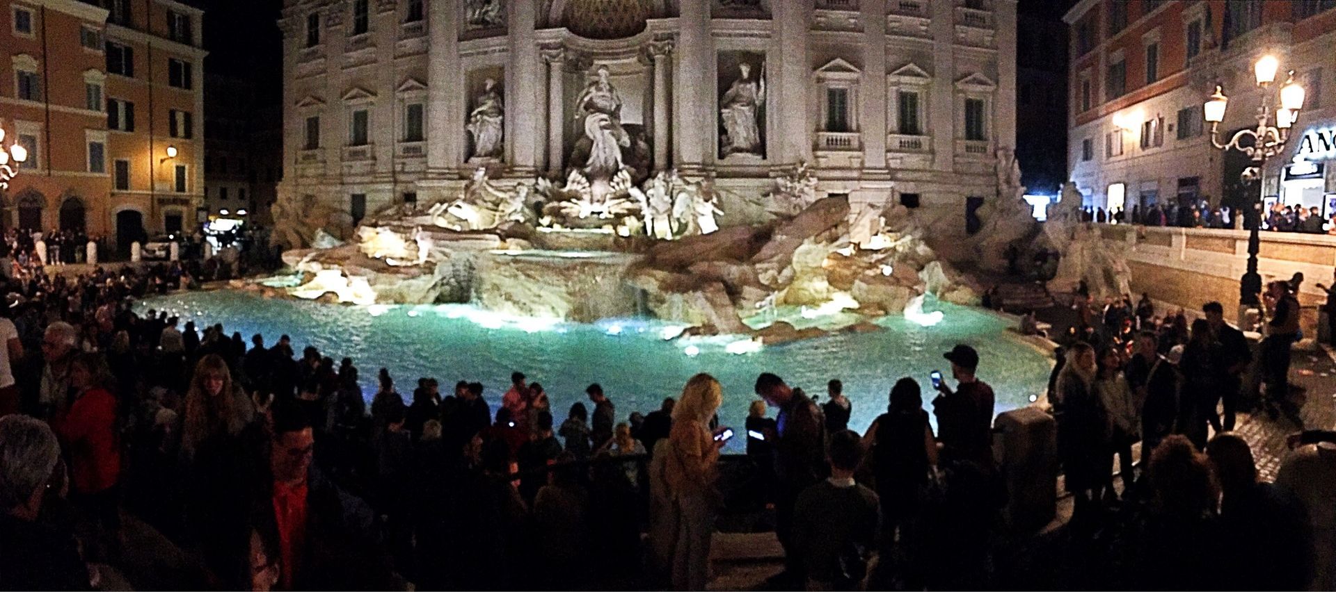 Crowd gathered at a lit fountain in a city square at night