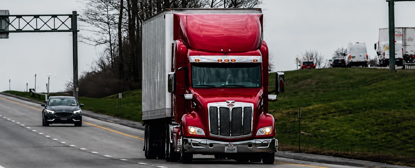 Red semi-truck driving on a highway beside a grassy embankment, with a car behind it.