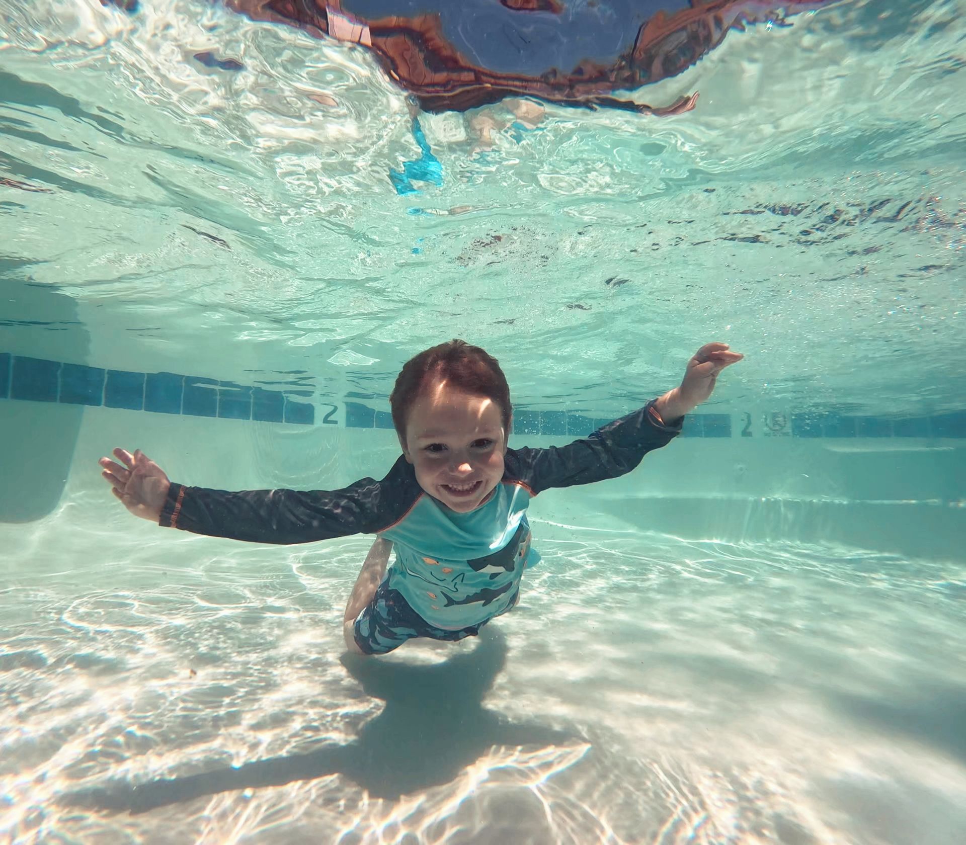 A child wearing a swim shirt smiles while swimming underwater in a bright, clear pool.