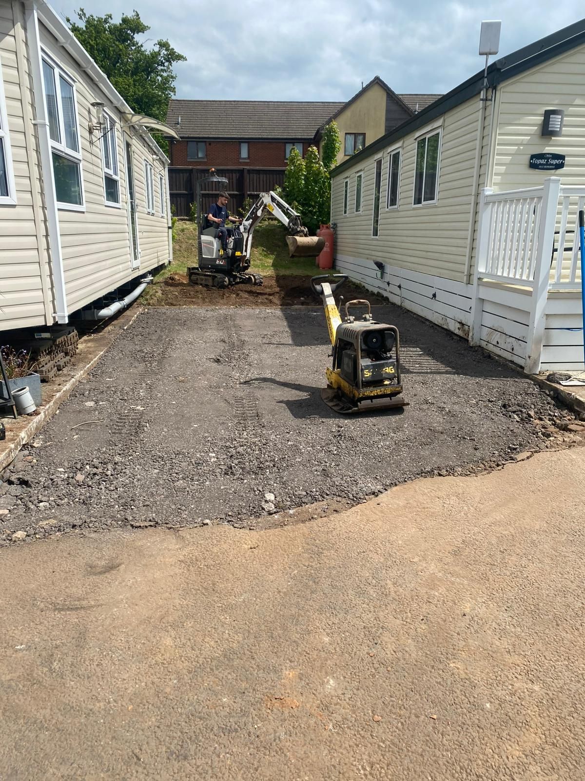 Construction site: mini-excavator and compactor work between two mobile homes, laying gravel on a sunny day.