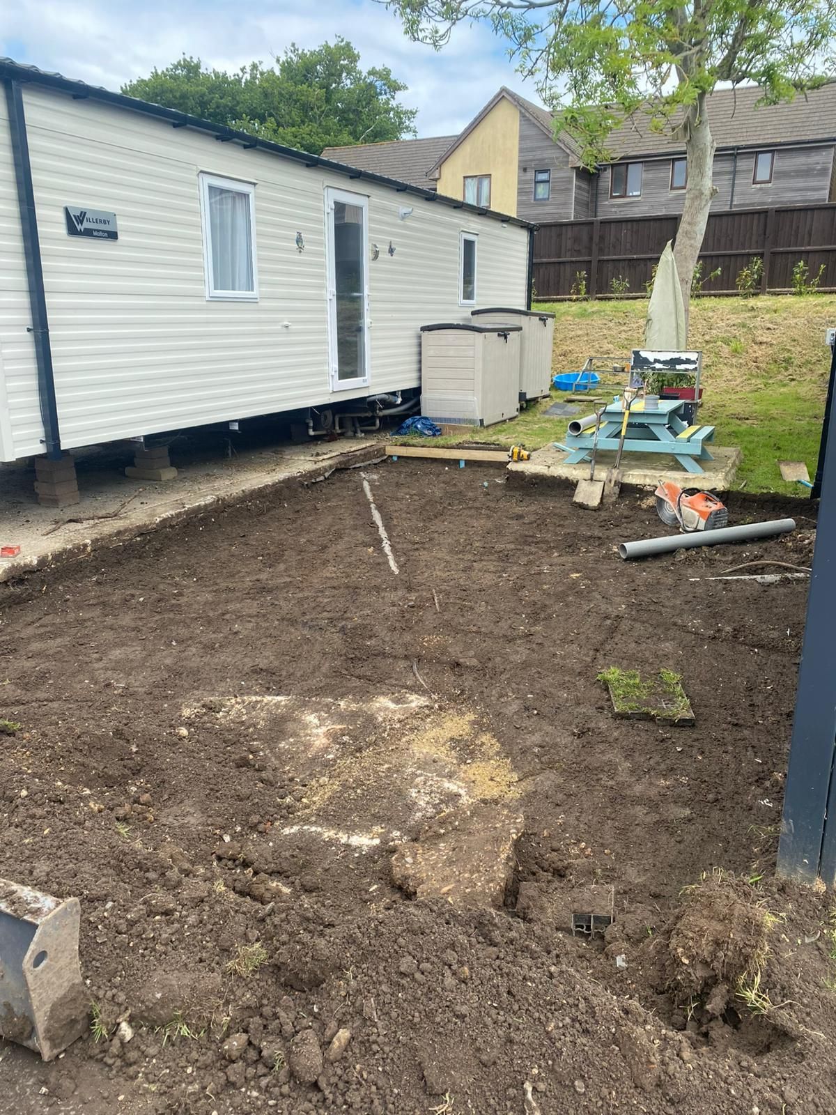 A mobile home with excavated ground, a picnic table, and trees in a yard.